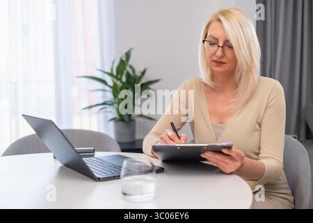 Psychologue féminine professionnelle offrant session en ligne dans un cadre chaleureux. Thérapeute blonde d'âge moyen dans des vêtements beiges à l'aide d'un ordinateur portable pour la consultation en ligne. Aide psychologique en ambiance conviviale. Banque D'Images