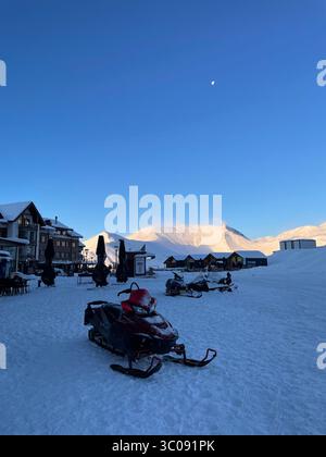 Une motoneige garée au fond d'une montagne enneigée dans une station de ski pittoresque. La lune est haute dans le ciel et le soleil sur le sommet de la montagne Banque D'Images