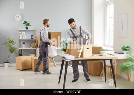 Déménagement de bureau, deux hommes en salopette emballant des boîtes, transportant des objets, plaçant des effets personnels sur le bureau Banque D'Images