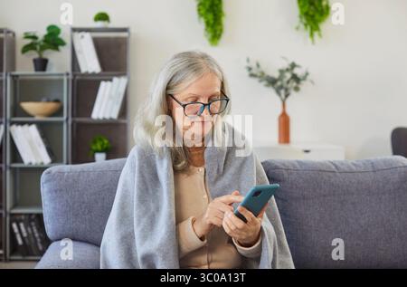 Femme senior enveloppée dans une couverture confortable assise sur le canapé t maison tenant le téléphone portable dans les mains. Banque D'Images