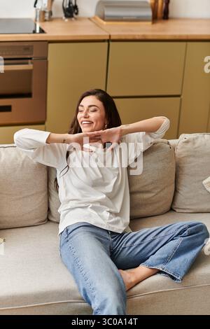 Une belle jeune femme aux cheveux bruns repose confortablement sur son canapé, souriante et détendue. Banque D'Images