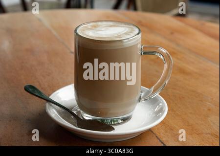 Tasse fraîchement infusée de cappuccino mousseux chaud avec garniture décorative de mousse de lait dans une grande tasse en verre sur une table en bois. Banque D'Images