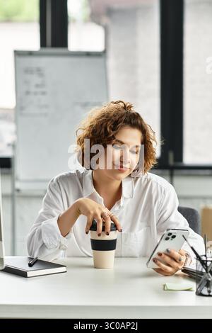 Femme d'affaires aux cheveux bouclés s'engage avec son smartphone tout en sirotant un café. Banque D'Images