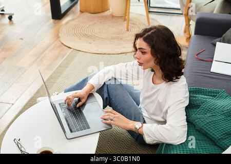 Jeune femme aux cheveux bouclés se concentre sur son ordinateur portable tout en profitant d'un espace de travail confortable à la maison. Banque D'Images