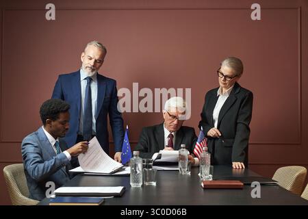 Homme caucasien d'âge moyen et homme caucasien senior assis à table, signant des documents avec une femme caucasienne senior et un homme noir d'âge moyen debout à proximité pendant la réunion politique Banque D'Images
