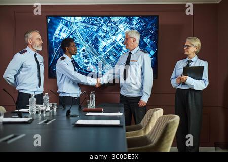 Homme caucasien d'âge moyen et homme noir d'âge moyen serrant la main dans la salle de conférence tandis que l'homme caucasien d'âge moyen et la femme caucasienne d'âge moyen observant, grand écran en arrière-plan Banque D'Images