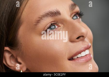 Une jeune femme rayonnante à la peau bronzée met en valeur son maquillage naturel sur un fond gris. Banque D'Images