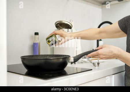 Une personne verse de l'huile d'olive dans une poêle placée sur une cuisinière électrique dans une cuisine Banque D'Images
