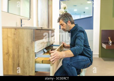 Homme mûr accroupissant, ouvrant les tiroirs d'une vanité de salle de bain, vérifiant la qualité et l'organisation dans une salle d'exposition de magasin d'amélioration de la maison Banque D'Images