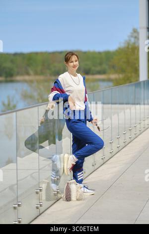 Une femme vêtue de vêtements de sport confortables se penche décontractée contre une balustrade en verre surplombant un plan d'eau serein Banque D'Images