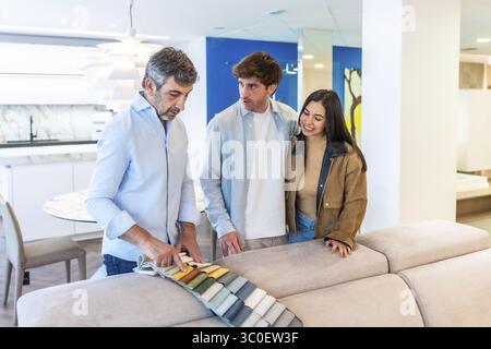 Couple regardant des échantillons de tissu avec le vendeur, choisissant la sellerie pour le nouveau canapé dans la salle d'exposition du magasin de meubles Banque D'Images
