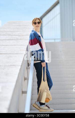 Une jeune femme sportive avec un sac à cordes se tient sur un escalier, habillée d'un survêtement athlétique élégant Banque D'Images