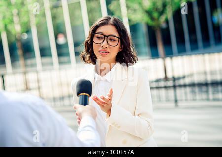 Femme d'affaires prospère interviewée à l'extérieur dans un cadre urbain par une journée ensoleillée, exprimant la confiance Banque D'Images