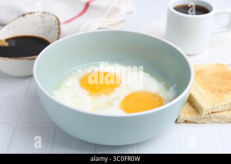 Petit déjeuner asiatique traditionnel avec œufs à moitié durs dans un bol, sauce soja, pain et café sur une table carrelée blanche, gros plan Banque D'Images