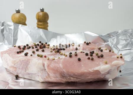 un morceau de viande de porc crue avec des pois de poivre sur papier d'aluminium est préparé pour la cuisson en gros plan Banque D'Images