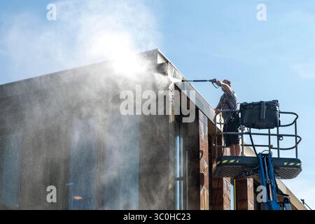 Homme debout sur la plate-forme de travail aérienne ou cueilleur de cerises avec pistolet à eau à haute pression nettoyant la façade d'un bâtiment moderne avec jet d'eau contre s. Banque D'Images