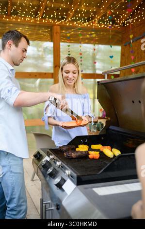 La compagnie joyeuse est occupée à préparer de la nourriture délicieuse sur le grill sur la véranda le jour d'été, profitant de l'atmosphère. Partie barbecue dans la maison de campagne. Amis au BBQ. Banque D'Images
