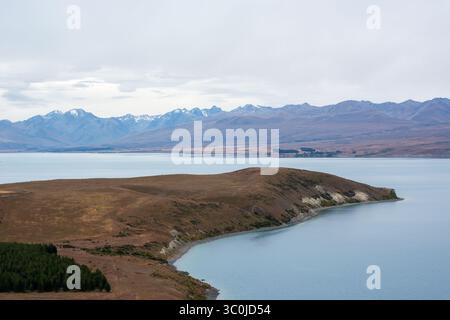 Un paysage serein avec un lac calme bordé par une colline en pente douce et des montagnes lointaines sous un ciel nuageux. Banque D'Images