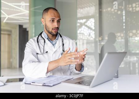 Un médecin, portant une blouse de laboratoire et un stéthoscope, fait des gestes pendant un appel vidéo à l'aide d'un ordinateur portable dans un environnement de bureau moderne. Banque D'Images