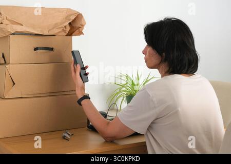 Femme vérifiant le téléphone tout en étant assis au bureau avec des boîtes à colis. Propriétaire de petite entreprise gérant des commandes en ligne ou recevant des livraisons. E-commerce Banque D'Images