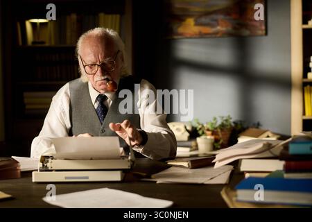 Homme caucasien senior avec des cheveux et des lunettes assis au bureau à l'aide d'une machine à écrire, tenant la pipe dans la bouche, entouré de papiers et de livres, apparaissant concentré sur l'écriture Banque D'Images