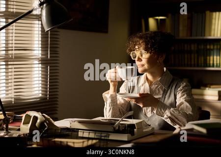 Femme caucasienne d'âge moyen assise à un bureau tenant une tasse et une soucoupe, portant des lunettes, regardant par la fenêtre, entourée d'étagères et d'équipements de bureau vintage, semblant réfléchie Banque D'Images