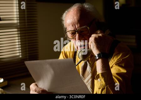 Homme caucasien senior parlant sur un téléphone fixe tout en lisant un document, portant des lunettes et une cravate, assis à l'intérieur près de la fenêtre, se concentrant sur la conversation et la paperasse Banque D'Images