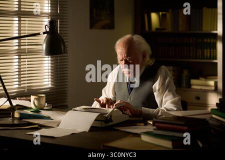 Homme caucasien senior assis au bureau tapant sur la machine à écrire, se concentrant sur l'écriture avec des papiers et des livres dispersés autour, la lumière du soleil coulant à travers les stores de fenêtre en arrière-plan Banque D'Images