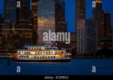 Jersey City, NJ - US - Jul 19, 2025 Un bateau à aubes classique glisse le long de la rivière Hudson à l'heure bleue, ses lumières s'échouent comme le skyli Lower Manhattan Banque D'Images