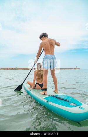 Jeune homme pagayant une planche gonflable avec une jeune femme assise dessus, profitant d'une journée ensoleillée à la plage Banque D'Images