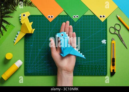 La main tient un jouet dinosaure origami fini en papier. Espace de travail pour l'art du papier avec des feuilles de papier colorées, des yeux googly, bâton de colle, stylo Banque D'Images