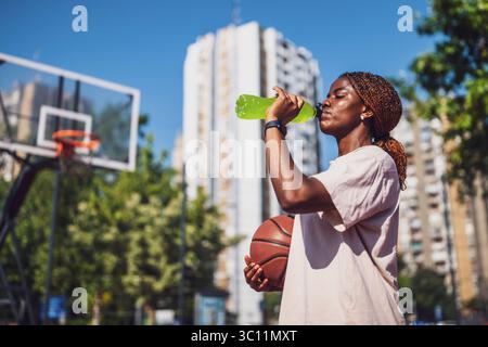 Jeune femme noire pratiquant le basket seule au terrain de sport le matin. Elle boit une boisson sportive. Banque D'Images