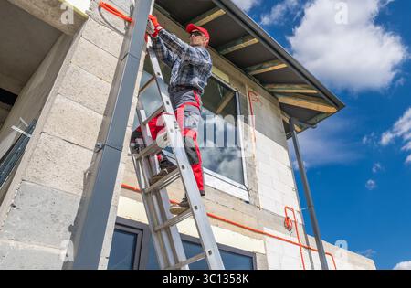 Un ouvrier de la construction dans une chemise à carreaux et un pantalon rouge monte une échelle pour installer des matériaux sur un bâtiment sous un ciel bleu avec des nuages moelleux. Banque D'Images