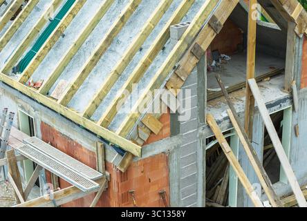 La progression de la construction montre des poutres en bois installées sur le nouveau toit d'une maison dans un cadre urbain, avec des matériaux de construction dispersés autour du site. Banque D'Images