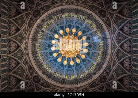 Muscat, Oman - 12 avril 2022 : vue sur le lustre rayonnant suspendu sous le dôme au design complexe, projetant une lueur chaude au milieu du plafond frais et à motifs. Banque D'Images