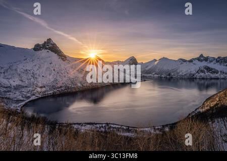 Vue aérienne du soleil éclatant sur les montagnes enneigées reflétées dans les eaux calmes du fjord, créant un paysage serein et contrasté, Senja, Troms og Finnmark, Norvège. Banque D'Images