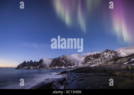 Vue sur les aurores boréales danses au-dessus de pics déchiquetés et enneigés reflétés dans l'eau tranquille, une symphonie de lumière et d'ombre, Troms, Norvège. Banque D'Images