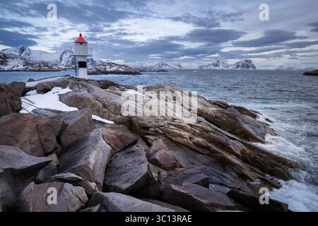 Vue d'un phare debout de garde sur des rivages rocheux sur fond de montagnes enneigées et de ciel sombre, Sørvågen, Norvège. Banque D'Images
