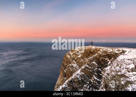 Vue sur le monument du globe debout au sommet des falaises poussiéreuses de neige, où l'océan sombre rencontre le ciel pastel doux, Nordkapp, Finnmark, Norvège. Banque D'Images