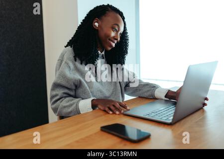 Une femme africaine engagée dans un rendez-vous virtuel, souriant tout en utilisant son ordinateur portable dans un cadre confortable à la maison. Elle porte des écouteurs, indiquant un foyer o Banque D'Images