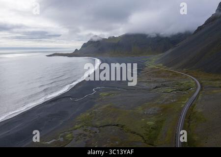 Vue aérienne d'une plage de sable noir embrassée par le bord de l'océan, ombragée par de majestueuses montagnes couvertes de brume, à côté d'une route sinueuse, Selfoss, Sveitarfélagið Árborg, Islande. Banque D'Images