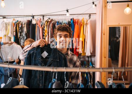 Un jeune homme tenant en toute confiance une veste élégante alors qu'il est à l'intérieur d'un magasin de vêtements tendance Banque D'Images