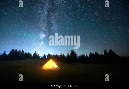 Tente touristique jaune incandescente sur une colline herbeuse sous un ciel étoilé époustouflant avec la voie lactée bien visible. Silhouette de pins et de montagnes. Beauté sereine de la nuit claire dans la nature. Banque D'Images