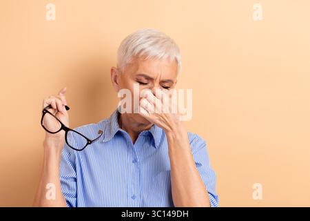 Portrait d'une femme mature tenant des lunettes et se pinçant le nez dans la frustration sur fond beige Banque D'Images