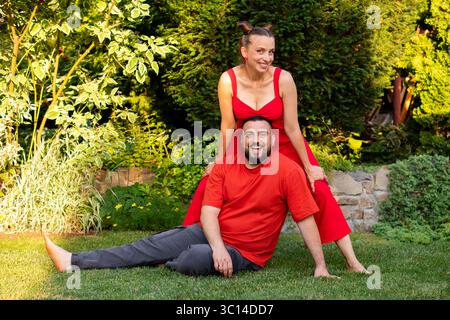 Une femme dans une tenue rouge s'appuie sur un homme assis portant du rouge dans un jardin luxuriant avec un feuillage vert et des fleurs en fleurs sous la lumière chaude du soleil Banque D'Images