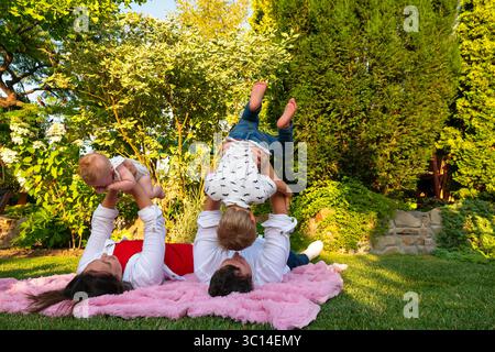 Couchés sur le dos, maman et papa soulèvent chaque enfant dans les airs au-dessus d'un tapis rose, créant des poses ludiques en miroir contre des arbustes luxuriants. Banque D'Images