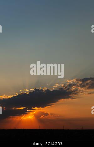 Un coucher de soleil doré brille avec des rayons lumineux du soleil perçant à travers des nuages sombres au-dessus d'un paysage lointain silhouette avec des poteaux électriques Banque D'Images
