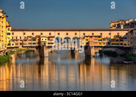 Vue sur les teintes dorées du Ponte Vecchio qui se reflètent sur le paisible fleuve Arno sous un ciel clair, créant une scène intemporelle, Florence, Toscane, Italie. Banque D'Images