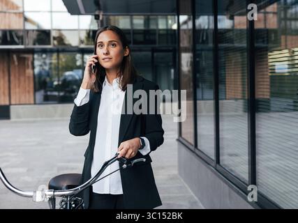 Jeune femme élégante dans des vêtements formels noirs avec vélo jeune femme élégante dans des vêtements formels noirs avec vélo Copyright : xxArtemxVarnitsinx Banque D'Images