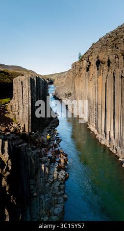 Niché en Islande, Studlagil Canyon présente des colonnes de basalte époustouflantes flanquant une rivière cristalline. Les visiteurs explorent cette merveille géologique sous un ciel bleu vif, mettant en valeur la beauté de la nature. Banque D'Images